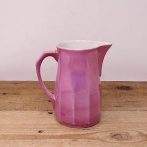 May include: A pink ceramic pitcher with a white interior. The pitcher has a handle and a spout. The exterior has a ribbed design. The pitcher is sitting on a wooden surface.