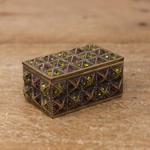 May include: A small, rectangular box with a decorative, geometric pattern. The box is made of metal and has a dark brown finish. The pattern is made up of small, green and brown rhinestones.