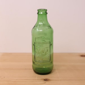 May include: A vintage green glass 7up soda bottle with a narrow neck and a rounded base. The bottle features embossed text that reads 7up, You Like It It Likes You, and No Deposit No Return, with a rectangular graphic frame around the brand name.