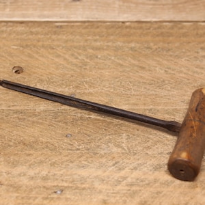 May include: A vintage wooden handled drill bit with a metal shaft. The drill bit is lying on a wooden surface.
