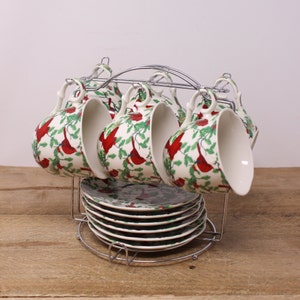 May include: Set of six white ceramic teacups and saucers featuring a red cardinal and green holly design. The cups and saucers are displayed on a chrome stand.