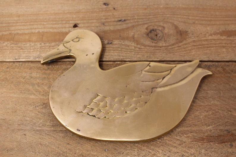 Pode incluir: Um descanso de panela de metal dourado em forma de pato. O pato tem um design detalhado de penas em suas asas.