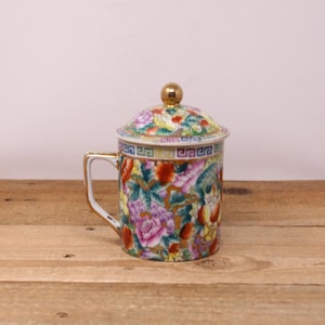 May include: A white porcelain teacup with a lid, featuring a colourful floral pattern with gold accents. The cup has a handle and a gold rim.
