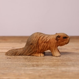 May include: A hand-carved wooden figurine of a beaver in a natural light brown finish. The sculpture features visible chisel marks across its body, a flat tail, and a defined head with small ears and dark eyes, capturing the animal in a low stance.