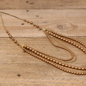 May include: A multi-strand gold necklace with brown faux pearl beads. The necklace is layered with different lengths of chain and beads.