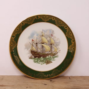 May include: A decorative plate featuring a detailed illustration of a sailing ship on a stormy sea. The plate has a white center with a green border and gold filigree accents. The ship has billowing sails and is set against a cloudy sky.