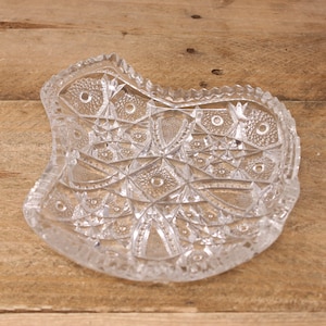 May include: Clear, decorative glass dish with a unique, asymmetrical shape. The dish features a detailed, cut-glass pattern with geometric shapes and a textured rim. The clear glass allows light to pass through, highlighting the intricate design.