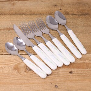 May include: A set of eight silver-toned cutlery pieces with white handles featuring a floral design. The set includes four forks, two spoons, a knife, and a butter knife.