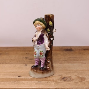 May include: A vintage porcelain figurine of a young person in a green hat, purple vest, and light blue pants with floral designs. The figurine is leaning against a brown tree trunk with green leaves. The base is a light brown color.