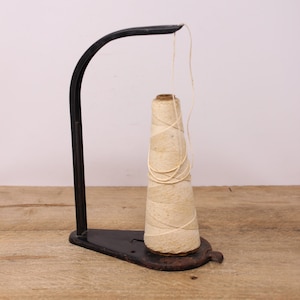 May include: A black metal thread stand with a spool of white thread. The stand has a curved arm and a base with a hole for the thread to pass through.