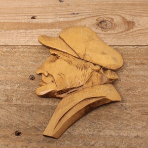 May include: A carved wooden wall hanging showing a man's profile wearing a cap. The artwork is light brown and features the name "BERTHIER" carved into the wood. The piece is mounted on a wooden surface, with visible wood grain.