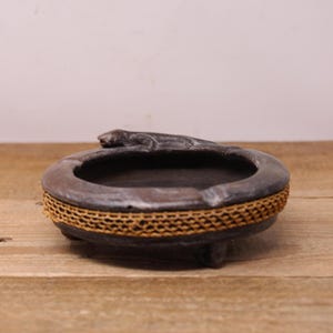 May include: A dark brown ceramic ashtray with a woven straw rim and a lizard-shaped handle. The ashtray is sitting on a wooden surface.