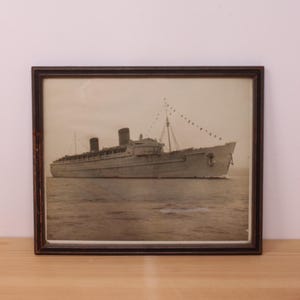 May include: A framed sepia-toned photograph of a large ocean liner sailing on the water. The ship features two dark smokestacks, multiple decks, and a mast with a string of signal flags extending toward the bow. The frame is dark brown wood.