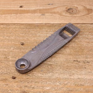 May include: A silver bottle opener with a textured design. The opener has a circular hole at one end and a bottle cap opener at the other. The body of the opener has a series of ridges and grooves.