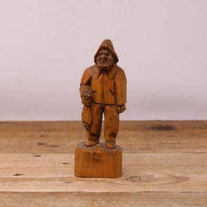 May include: A hand-carved wooden figurine of a fisherman, wearing a hat and holding a fish. The figure is standing on a rectangular base. The wood has a warm, natural tone.