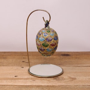May include: A decorative egg-shaped ornament with a gold filigree design, featuring blue, green, and purple accents. The ornament hangs from a curved, gold-coloured metal stand with a circular mirror base. The egg is approximately 7.6 cm tall.