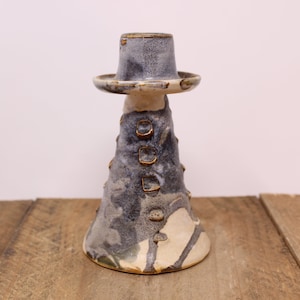 May include: A handcrafted ceramic candlestick holder with a unique, textured design. The candlestick features a dark blue and cream colour scheme with gold accents. The base has a flared shape, and the top has a small, hat-like design.
