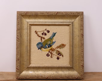 May include: A framed needlepoint artwork featuring a blue, yellow, and green bird perched on a branch with berries. The bird is set against a beige background and framed in a gold-colored, ornate wooden frame.