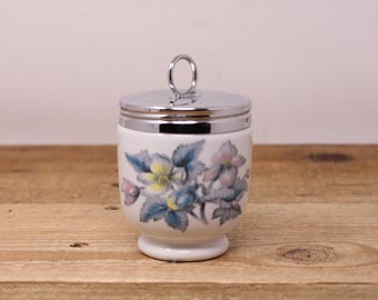 May include: A white ceramic egg cup with a floral design in blue, yellow, and pink. The egg cup has a silver-colored metal lid with a small handle. The cup sits on a wooden surface.