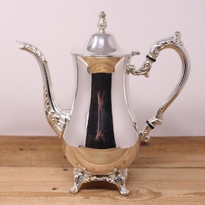 May include: A polished silver teapot with decorative flourishes. The teapot has a curved spout, an ornate handle, and a lid with a small finial. It stands on a three-footed base, and the shiny surface reflects the surroundings.