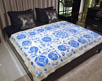 Puede incluir: Una cama con una colcha blanca y azul con estampado floral. La colcha es el punto focal, con un diseño detallado de flores y hojas azules sobre un fondo blanco. Almohadas negras y un marco de cama oscuro completan la escena.