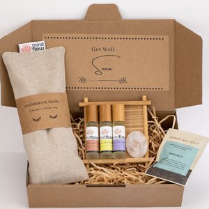 May include: A brown cardboard box filled with a self-care gift set. The box contains a lavender eye pillow, three essential oil roller bottles, a wooden comb, a rose quartz crystal, and a bag of Byron Bay Tea Co. Immunity tea. The box has a card on top that says "Get Well Soon".