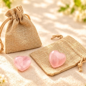 May include: Two smooth, polished, light pink heart-shaped stones made of quartz. One stone rests on a small, tan, woven burlap drawstring pouch, while the other stone sits beside a second, upright, matching burlap drawstring pouch.