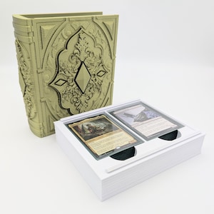 May include: A sage green, book-shaped card holder with ornate detailing and a diamond-shaped window. A white card case holds two trading cards. The holder is designed to resemble a vintage book.