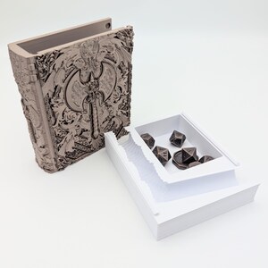 May include: A detailed brown book-shaped box featuring an axe design. A white tray inside holds bronze polyhedral dice. The box is open, displaying the dice and a stack of white paper. The box is likely for storing dice.
