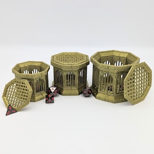 May include: Three 3D printed, gold-colored, hexagonal cages with a lattice design. The cages are open on top and have a removable lid. The largest cage has its lid off, revealing the interior.  There are five black and red dice on the table.