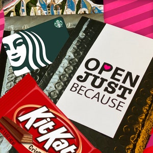 Może przedstawiać: Biała kartka z tekstem "OPEN JUST BECAUSE" w kolorze czarnym z r&oacute;żowym symbolem serca. Kartka leży na czarno-srebrnym tle z folii bąbelkowej. Widoczne są r&oacute;wnież zielono-biała karta podarunkowa Starbucks i czerwono-biały baton Kit Kat.
