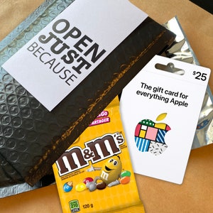 Może przedstawiać: Czarno-biała karta podarunkowa z tekstem "The gift card for everything Apple" i kolorowym wzorem jabłka. Karta podarunkowa jest warta 25 $. Karta podarunkowa leży na brązowej kopercie bąbelkowej z tekstem "OPEN JUST BECAUSE" na białym kartonie.