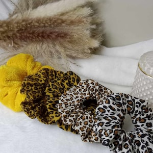 Peut inclure: Cinq chouchous de cheveux dans une variété de motifs. L'un est jaune uni, et les quatre autres sont à imprimé léopard dans différentes combinaisons de couleurs. Les chouchous sont disposés en rangée sur une surface blanche.