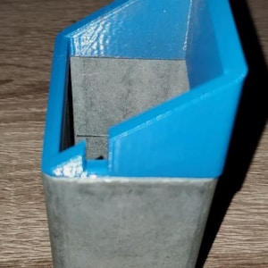 May include: A two-tone container with a blue top and a gray base. The top section is a vibrant blue, with a geometric, angular design. The base is a solid, rectangular shape. The item is likely 3D-printed.