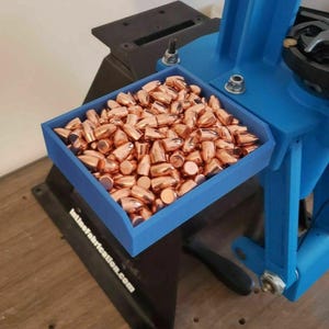 May include: A blue tray filled with copper-colored bullets sits next to a blue reloading press. The tray is rectangular and holds numerous bullets. The reloading press is partially visible, with a black base and various mechanical components.