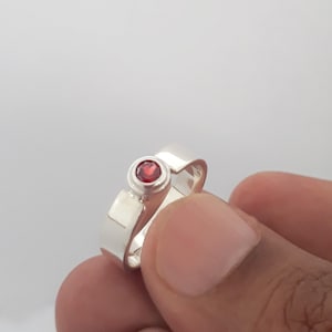 May include: A silver ring with a red gemstone set in the center. The ring is open and adjustable.