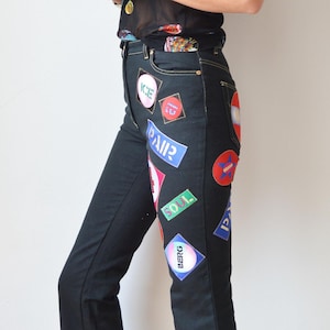 May include: Black denim jeans with a variety of colourful patches, including words like "ICE", "SOUL", and "BERKS". The jeans have a bootcut style and are paired with black platform shoes.
