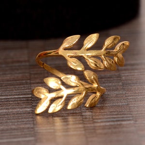 May include: A gold-coloured ring with a leaf design. The ring wraps around the finger with two branches of leaves. The leaves have detailed veining. The ring is set against a wooden surface.