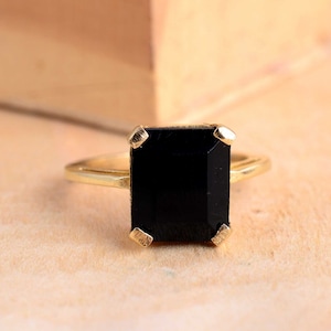 Puede incluir: Anillo de oro con una gran piedra negra rectangular. La piedra está sujeta por cuatro garras de oro. La banda del anillo es de oro liso. El anillo está sobre un fondo neutro. Joya de estilo clásico.