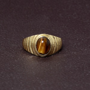 May include: A gold ring featuring an oval tiger's eye gemstone. The ring has a textured band with vertical lines and a polished finish. The tiger's eye stone is brown with golden streaks, set in a smooth bezel.