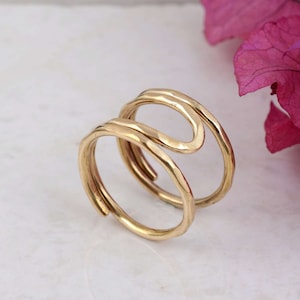 May include: A gold-colored ring with a unique, flowing design. The ring features a double-banded, open design with a hammered texture. The ring is set against a white background with a pink flower in the corner. A stylish piece of jewelry.