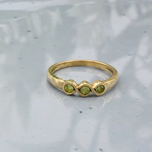 May include: A gold ring featuring three round, light green gemstones set in a row. The ring has a simple, elegant design with a polished finish. The gemstones are evenly spaced and add a pop of color to the gold band. Jewelry.