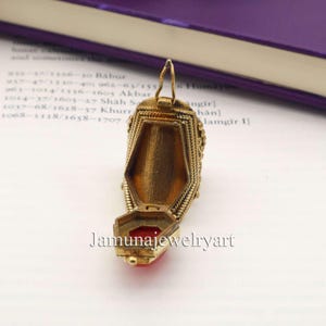 May include: A gold-colored, ornate pendant with an open compartment revealing a red interior. The pendant has intricate detailing and a hinged lid. The text "Jamunajewelryart" is visible on the image.
