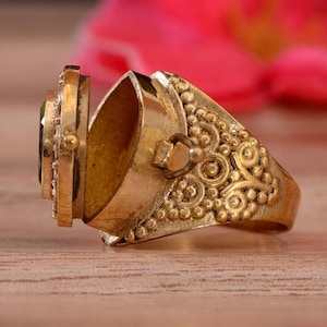 May include: A gold-colored locket ring with an oval-shaped compartment. The ring features an ornate design with a green gemstone and intricate detailing. The ring is open, revealing the hollow interior.
