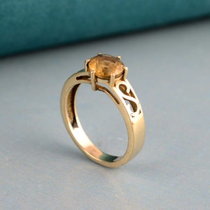 May include: A gold ring with a round, light orange gemstone. The ring has a decorative openwork design on the sides of the band. The gemstone is held in place by four prongs. The ring is set against a light blue background.