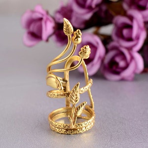 May include: A gold-colored finger ring with an intricate floral design. The ring features a base with a detailed pattern, multiple bands, and delicate leaf and bud accents. The ring is set against a soft background with purple flowers.