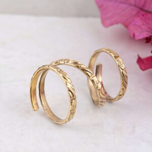 May include: A gold-colored double finger ring with floral and vine detailing. The ring wraps around two fingers, with a connecting band featuring similar embossed patterns. The design is intricate and the ring has a polished finish.