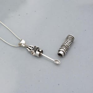 May include: A silver-toned metal pendant necklace featuring a detachable cylindrical cap with intricate rope-like textures. The pendant base includes a small, thin spoon-like attachment extending outward, connected to a delicate silver chain link necklace.