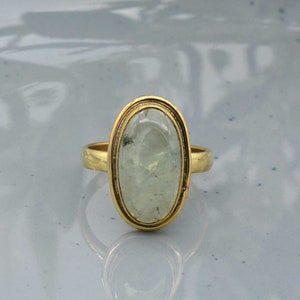 以下が含まれることがあります： 楕円形の淡い緑色の宝石をあしらったゴールドリング。 宝石は金のベゼルにセットされています。 リングのバンドはシンプルな磨かれたゴールドです。 宝石はわずかに半透明です。