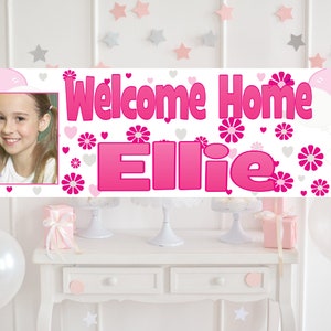 Puede incluir: Una pancarta de bienvenida a casa rosa y blanca con una foto de una ni&ntilde;a y el nombre "Ellie" en letras rosas. La pancarta est&aacute; decorada con corazones y flores rosas.